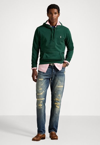 Groene hoodie met een voorzak, gelaagd over een roze shirt. Draagt het met vervaagde blauwe jeans met versleten details. Bruine nette schoenen.