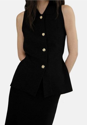 Blusa negra sin mangas con botones dorados, usada sobre una falda negra a juego, con las manos del modelo detrás de la espalda sobre un fondo sencillo.