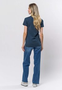 Tumši zaļa īsroku T-krekls, brīva piegriezuma, apvienots ar gaiši zilām plaša piegriezuma džinsām un baltām sporta kurpēm, skatīts no aizmugures.