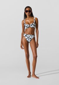 Bikini-sæt i sort og hvidt med bladmønster, der har en struktureret top med sløjfe foran og højtaljede bukser. Glat stof, brede stropper, minimal hardware.