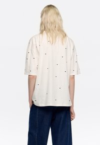 Biała bawełniana koszulka z czarnymi ćwiekami, luźny krój, krótki rękaw oraz rozcięcie po bokach u dołu, noszona z ciemnoniebieskimi, luźnymi dżinsami.