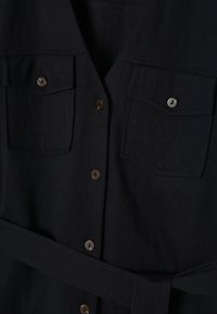 Robe chemise noire avec fermeture à boutons sur le devant, deux poches poitrine avec boutons et ceinture en tissu assorti à la taille.