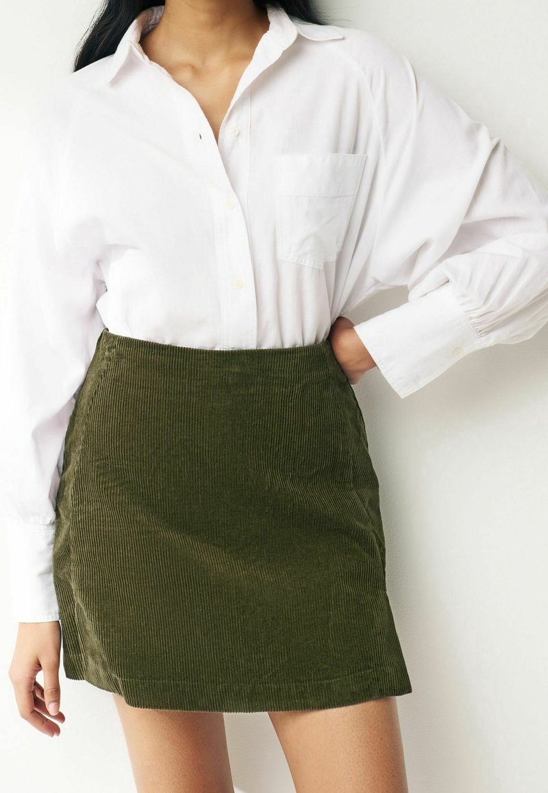 Camicia bianca con bottoni e tasca, abbinata a una gonna in velluto a coste verde. La gonna presenta un motivo a coste testurizzato e una linea dritta.
