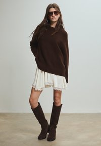 Maglione oversize marrone, gonna bianca con bordo in pizzo e stivali al ginocchio in suede marrone. Modella che indossa occhiali da sole grandi marroni, in piedi contro uno sfondo neutro.