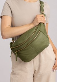 Donna vestita con abiti beige che tiene una borsa a tracolla in pelle verde oliva con due cerniere dorate sul petto.