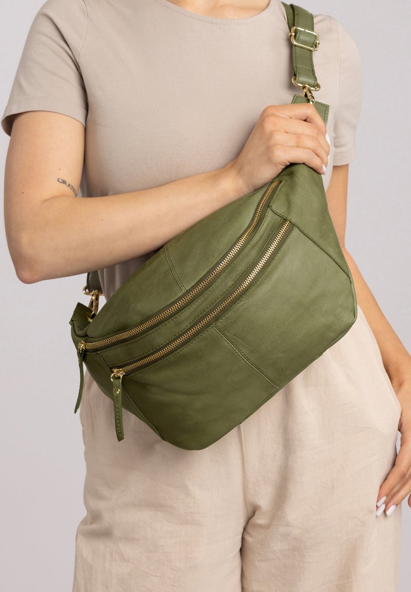 Donna vestita con abiti beige che tiene una borsa a tracolla in pelle verde oliva con due cerniere dorate sul petto.