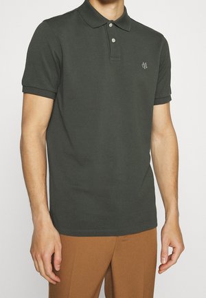 Mężczyzna ubrany w ciemnozieloną, krótko rękawową koszulkę polo z dwoma guzikami oraz brązowe spodnie na jednolitym tle.