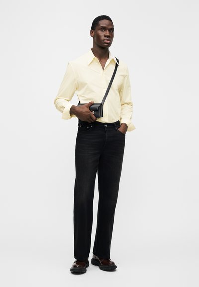 Jasnożółta koszula na guziki, czarne dżinsy, czarna torebka noszona na krzyż, brązowe buty. Model stoi pewnie z rękami w kieszeniach na jednolitym tle.