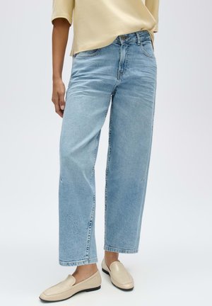 Persona che indossa jeans a gamba dritta azzurri chiari, mocassini beige e una maglietta giallo chiaro a maniche corte, in piedi davanti a uno sfondo semplice.