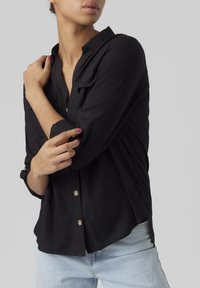 Camicia nera con bottoni e maniche arrotolate, realizzata in un tessuto liscio. Presenta due tasche sul petto e bottoni color oro. Indossata sopra jeans azzurri.