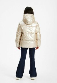 Vadderad, huva jacka i glänsande beige med en puffig design. Kombinerad med mörka utsvängda jeans, vilket visar en avslappnad vinteroutfit.