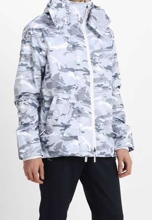 Person iført en jakke med lys grå og hvid camouflageprint, høj krave og lynlås foran, kombineret med sorte bukser.