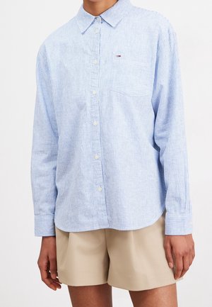 Person trägt ein hellblau-weiß gestreiftes Langarmhemd mit Knöpfen und beige Shorts vor schlichtem Hintergrund.