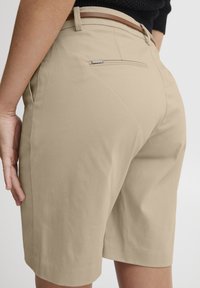 Beige skreddersydde shorts laget av glatt stoff, med en baklomme med metallisk detalj. Knelengde med et rent, strukturert design.