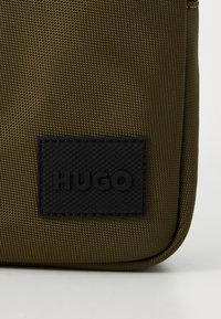 Oliven grønn tekstilpose med en strukturert svart gummilogo med teksten "HUGO." Har glidelås og forsterket søm langs kantene.