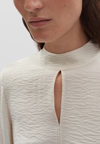 Primo piano della parte inferiore del viso e del collo di una donna che indossa una blusa di cremoso consistenza con colletto alto e un'apertura a forma di chiave verticale.