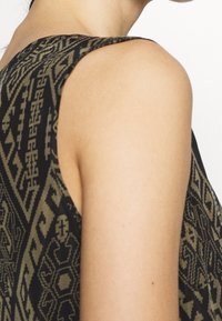 Axel och övre rygg på en person som bär en ärmlös topp med ett svart och olivgrönt geometriskt mönster på en vit bakgrund.