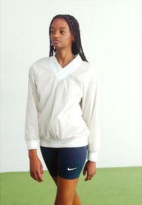 Ελαφρύ, ανοιχτό μπεζ πούλιερ με V-λαιμό, ελαστικές μανσέτες και ελαστική μέση, συνδυασμένο με ναυτικό σορτσάκι που διαθέτει λευκό λογότυπο. Απαλή υφή.