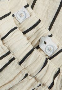 Ljust färgad, texturerad tyg med svarta ränder och vita knappdetaljer; har insydda sektioner för extra detalj.
