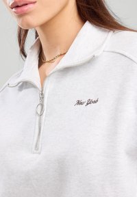 White zip-up pullover with a high collar, featuring a silver zipper and embroidered "New York" in dark lettering. Soft, textured fabric.