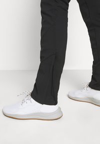 Pantalon de sport noir avec fermetures éclair latérales, fabriqué en matériau léger, accompagné de baskets blanches avec une semelle texturée et des lacets multicolores.
