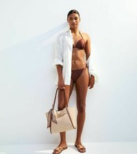 Brun bikini med trekantig topp och knytband i sidorna, kombinerad med en lös vit skjorta och en stor beige strandväska med ett mönstrat accent.