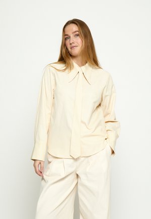 Ženska z dolgimi blond lasmi, oblečena v kremasto, preveliko srajco z dolgim ovratnikom in kremasto širokimi hlačami na belem ozadju.