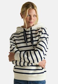 Młoda kobieta z blond włosami w granatowym i białym w paski hoodie, złożonymi rękami, patrząca na bok, na jednolitym tle.