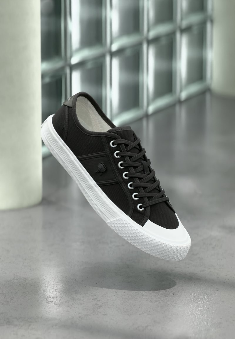 Μαύρο αθλητικό παπούτσι χαμηλού τύπου με λευκή λαστιχένια σόλα και πρόσθια κάλυψη, μαύρα κορδόνια και λογότυπο στο πλάι, αιωρούμενο πάνω από γκρι γυαλιστερό πάτωμα.