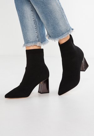 Personne portant des bottines chaussettes en tricot noir avec talons blocs associées à un jean bleu clair à ourlet effiloché sur fond blanc.