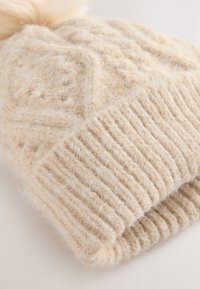 Knit beige beanie made from soft, textured wool. Features a ribbed cuff and a fluffy pom-pom on top, showcasing a geometric pattern.