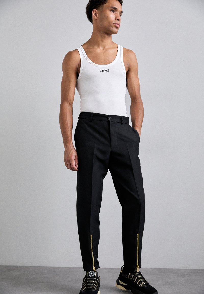 Zwarte op maat gemaakte broek met een ritsaccent bij de enkels, gecombineerd met een witte mouwloze tanktop met een logo, en zwarte sneakers.