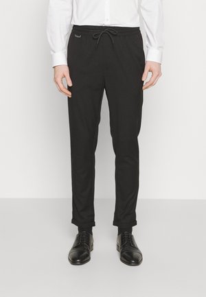 Black trousers with a drawstring waist, slim fit, and rolled cuffs. Made from smooth fabric; paired with polished black shoes.
