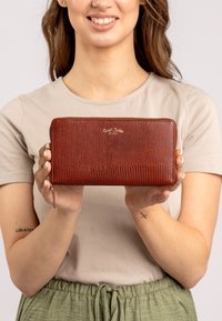 Raudona tekstūruota piniginė su užtrauktuku, puikuojanti auksiniu logotipu ir riešo dirželiu, laikoma rankose prieš neutralų foną.