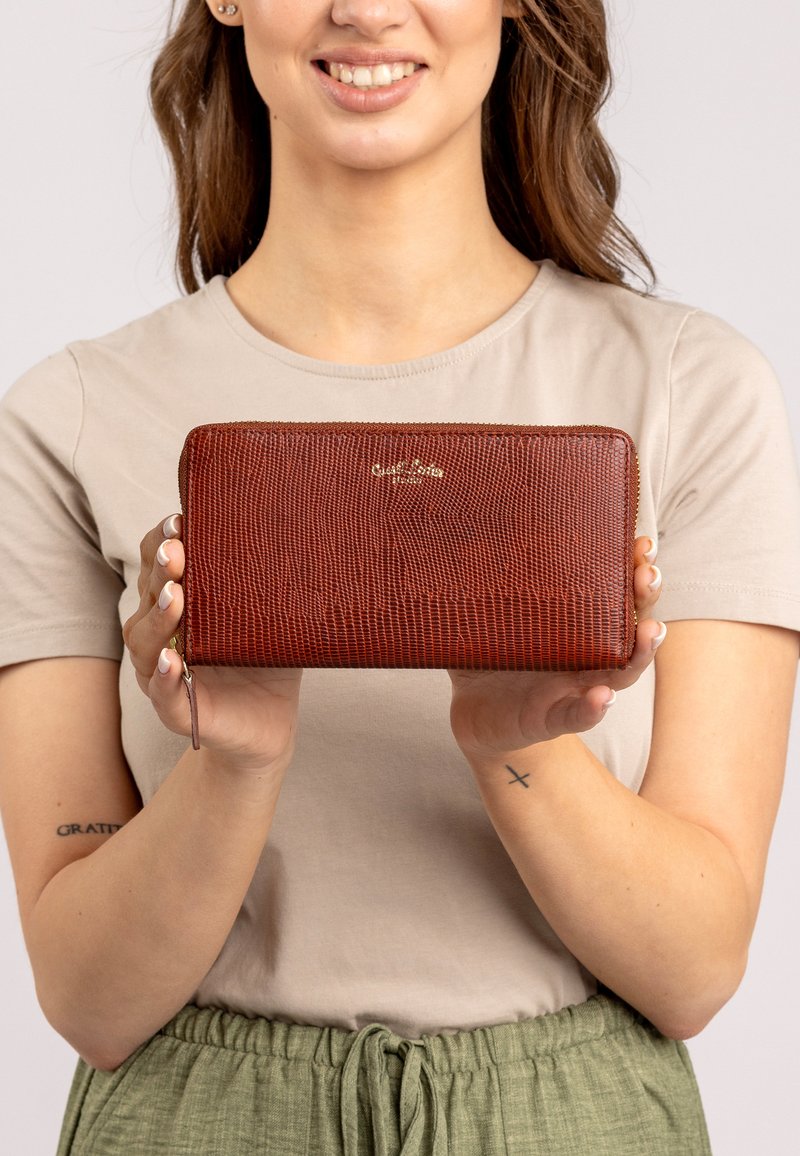 Raudona tekstūruota piniginė su užtrauktuku, puikuojanti auksiniu logotipu ir riešo dirželiu, laikoma rankose prieš neutralų foną.