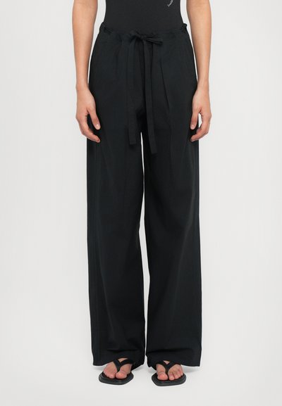 Pantaloni a gamba larga neri realizzati in tessuto leggero, con vita regolabile e tasche laterali. Indossati con ciabatte nere.