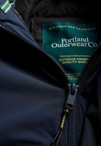 Granatowa kurtka outdoorowa z gładkiego materiału, z zapięciem na zamek z przodu oraz zieloną metką z napisem "Portland Outerwear Co." w złotym kolorze. 