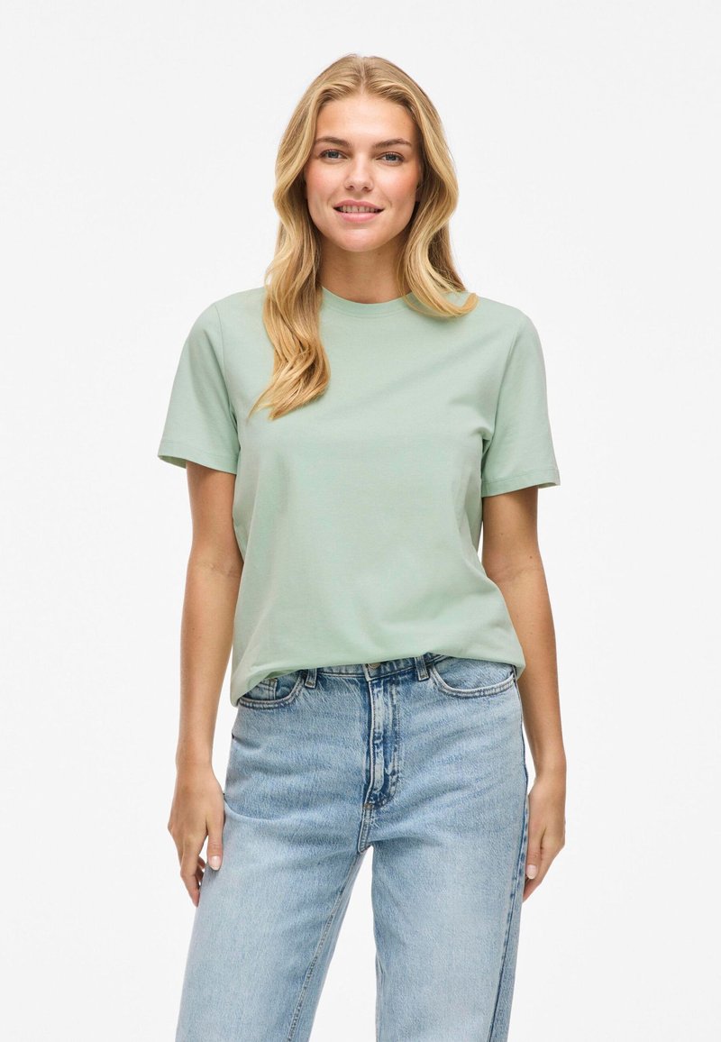 Gai zaļš kokvilnas T-krekls ar īsām piedurknēm, apaļu kakla izgriezumu, brīvu piegriezumu, valkāts ar augsto vidukļa zilu džinsu biksēm. Vienkāršs un klasiskš dizains.