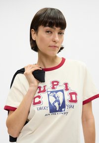 Βαμβακερή μπλούζα σε κρέμα με κόκκινες λεπτομέρειες, featuring μια γραφική εκτύπωση ενός χαρακτήρα και το κείμενο "CUPID LUCKY YOU" σε έντονα χρώματα.