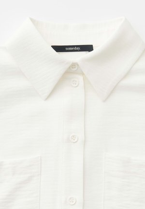 Wit overhemd met knopen, voorzien van een puntige kraag en twee borstzakken, met een zwart label in de kraag met de tekst "someday.".