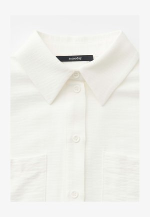 Chemise blanche à boutons avec un col pointu et deux poches poitrine, comportant une étiquette noire avec le texte « someday. » à l'intérieur du col.