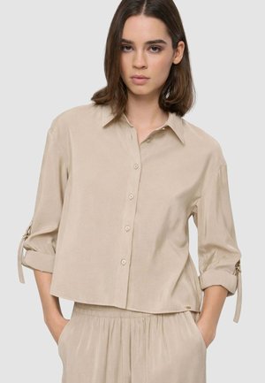 Donna che indossa una camicia beige con bottoni e maniche arrotolate e pantaloni larghi abbinati, in piedi con le mani nelle tasche davanti a uno sfondo semplice.