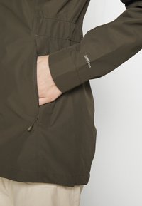 Main insérée dans la poche latérale zippée de la veste verte, la manche affiche le logo « DRYVENT », pantalon beige visible sous l'ourlet de la veste.