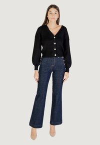 Cardigan nero a coste con scollatura a V e grandi bottoni, abbinato a jeans a zampa d'elefante blu scuro a vita alta con bottoni laterali.