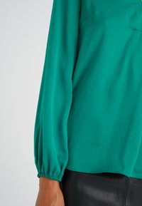 Blusa color teal con maniche lunghe e puffate, con polsini arricciati, realizzata in tessuto leggero. Texture morbida con una vestibilità rilassata.