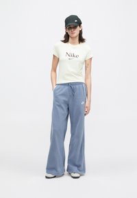 Mladá žena v čiernej čiapke Nike, krémovom tričku Nike, voľných modrých nohaviciach Nike a bielych teniskách stojí na pozadí bez vzoru v bielej farbe.