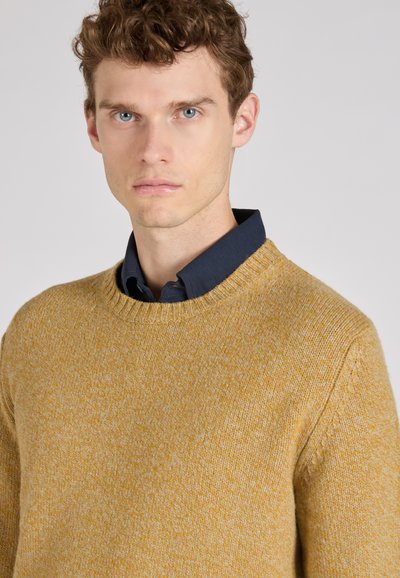 Pull en maille jaune moutarde avec un col rond et des poignets côtelés, superposé à une chemise bleu marine à col. Texture lisse, design décontracté.