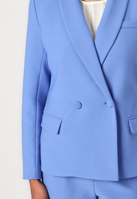 Lys blå dobbeltradet blazer laget av teksturert stoff, med skrå krage, to frontlommer og subtile knappedetaljer.