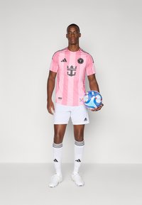Rosa fodboldtrøje med sorte accenter, hvide shorts og sokker med sorte striber. Spilleren holder en blå og hvid fodbold.