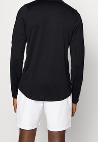 Musta pitkähihainen urheilu-toppi, jossa on pyöreä helma, ja jossa on sileä pinta sekä raglan-hihat. Yhdistetty valkoisiin shortseihin, mikä luo rentoa tyyliä.